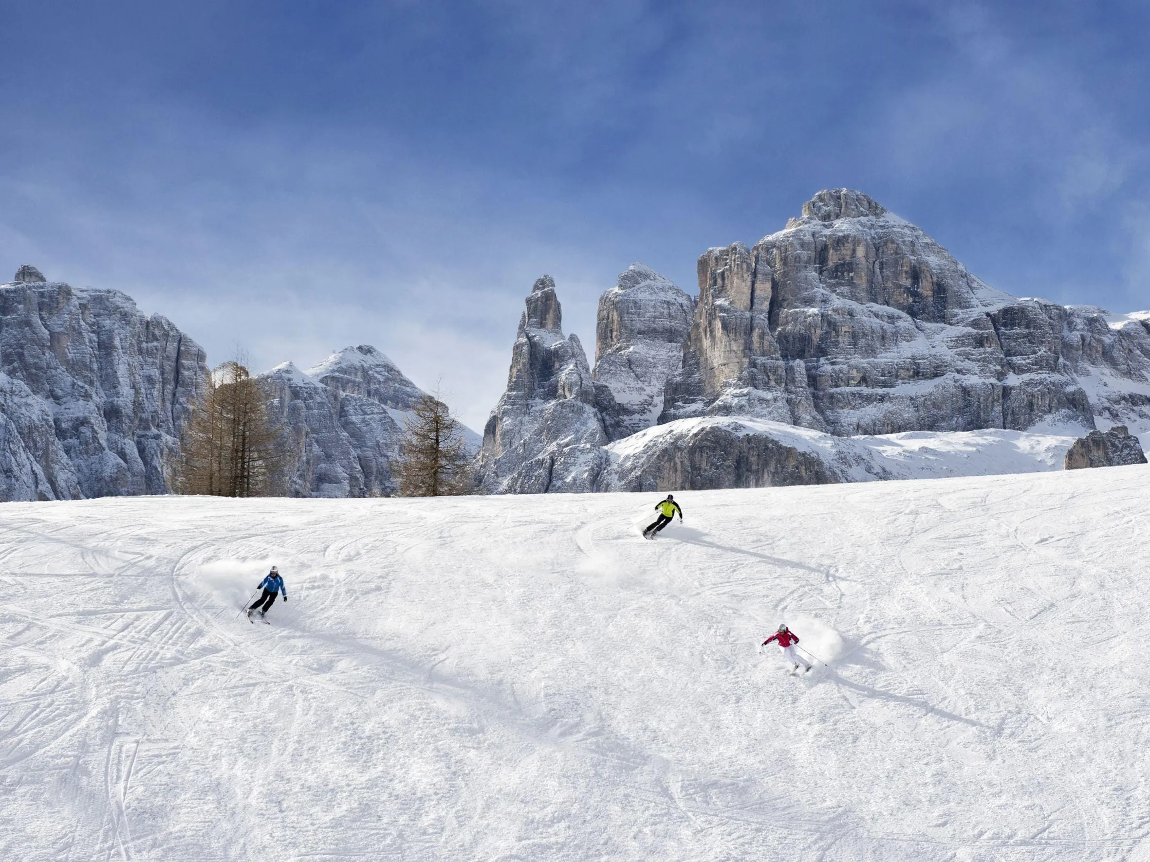 Three skiers navigate snowy slopes against a dramatic backdrop of tall, rugged Dolomites.