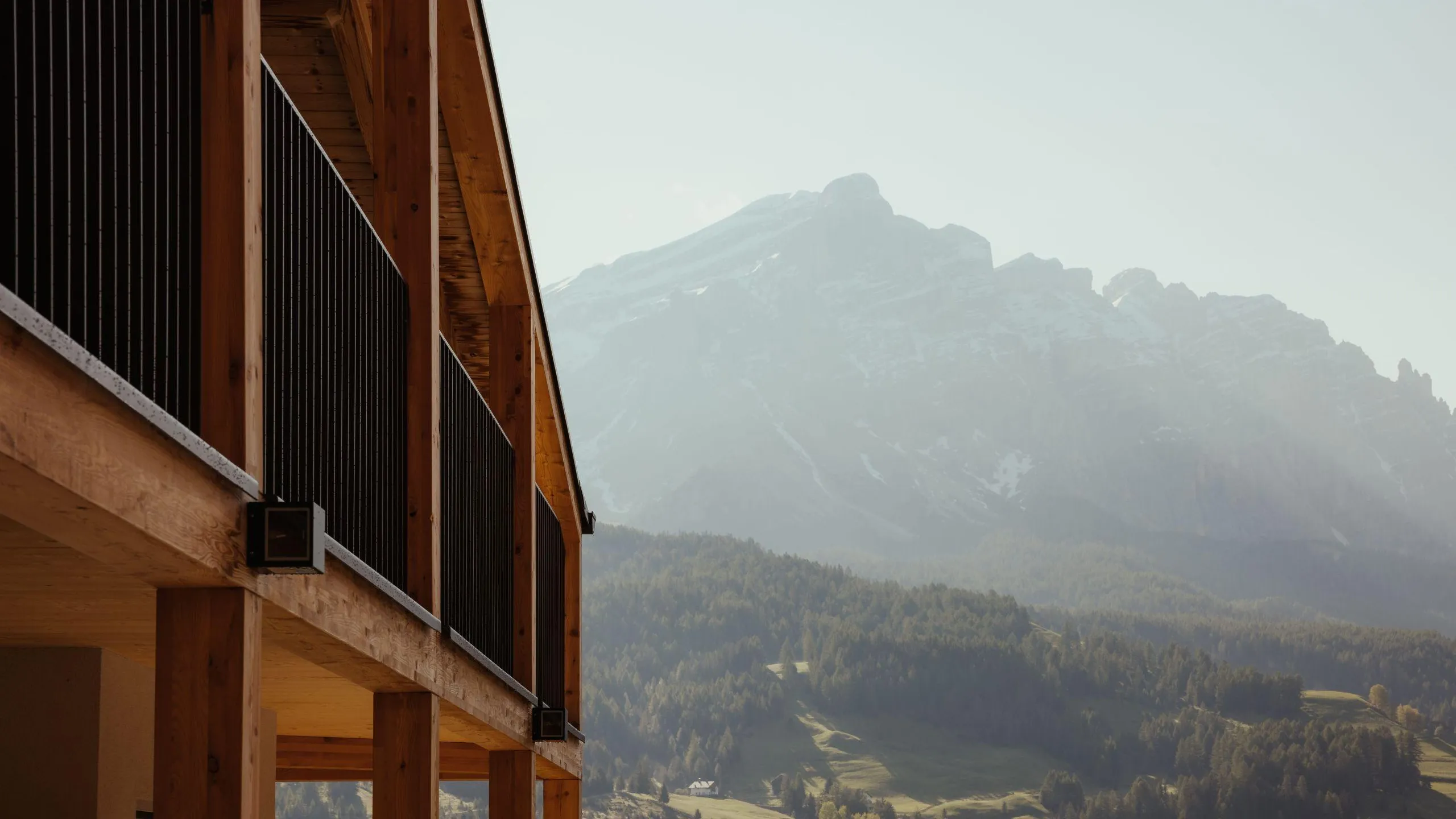 The wooden balcony of the Hotel Lech da Sompunt in La Villa frames the distant mountain range of La Varella, with lush green hills in the foreground and soft morning light creating a serene atmosphere.