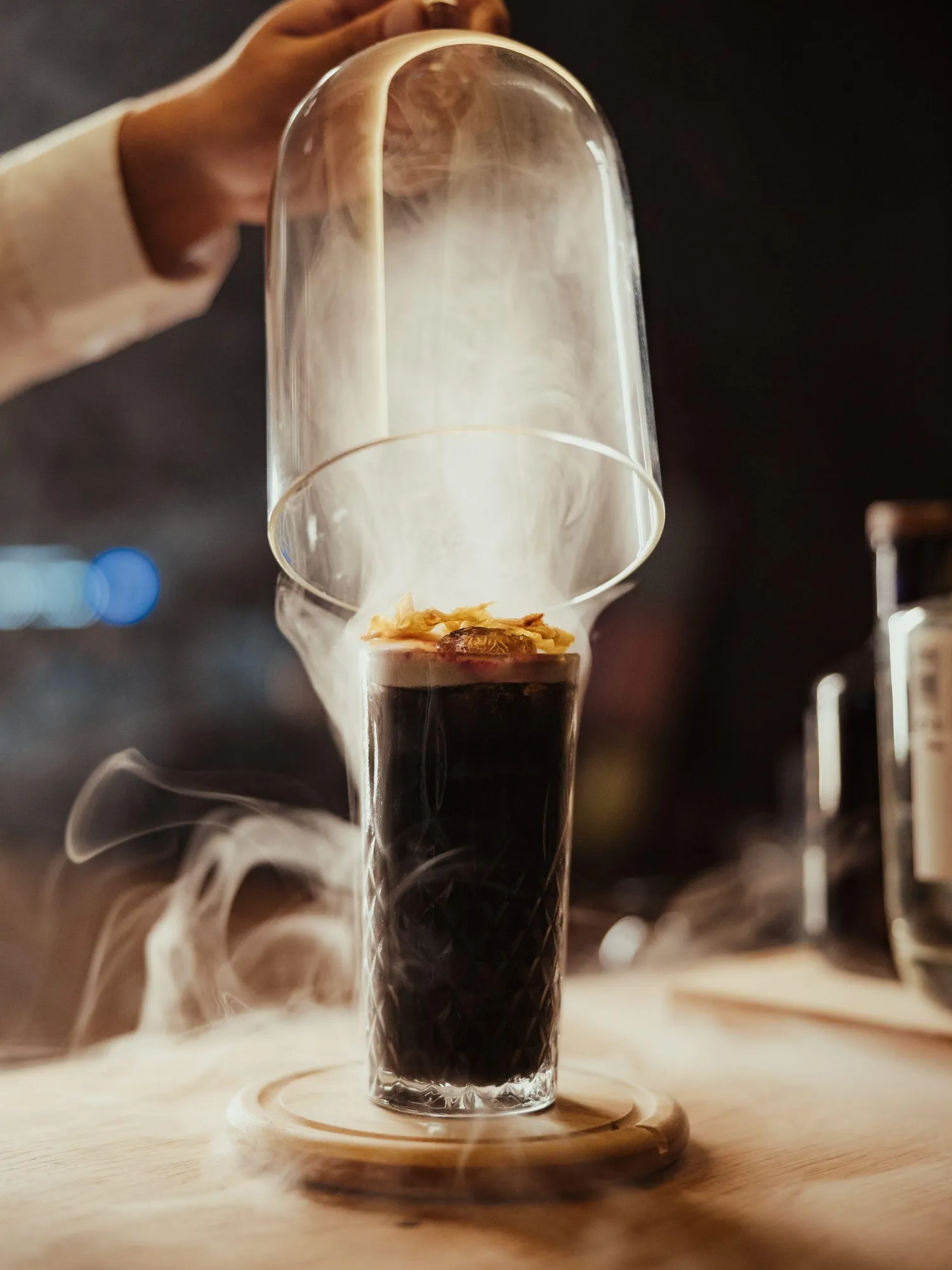 Una mano solleva una cupola di vetro, rivelando un cocktail fumoso guarnito con fiori secchi, posto su un tavolo di legno in un accogliente bar dell'Hotel Lech da Sompunt.
