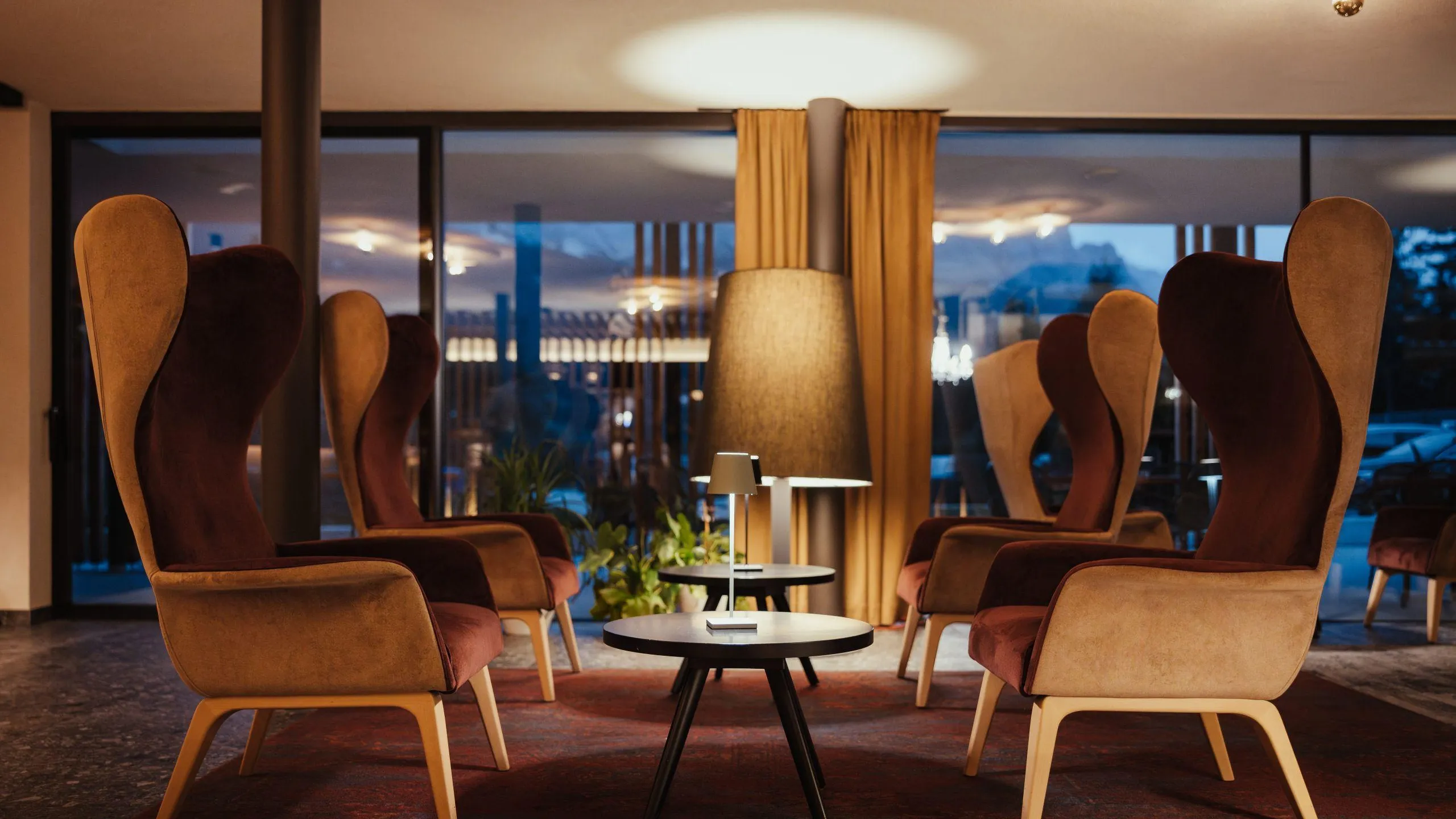 Cozy seating area in Hotel Lech da Sompunt, featuring stylish high-back chairs, low tables, and warm lighting against a backdrop of large windows.