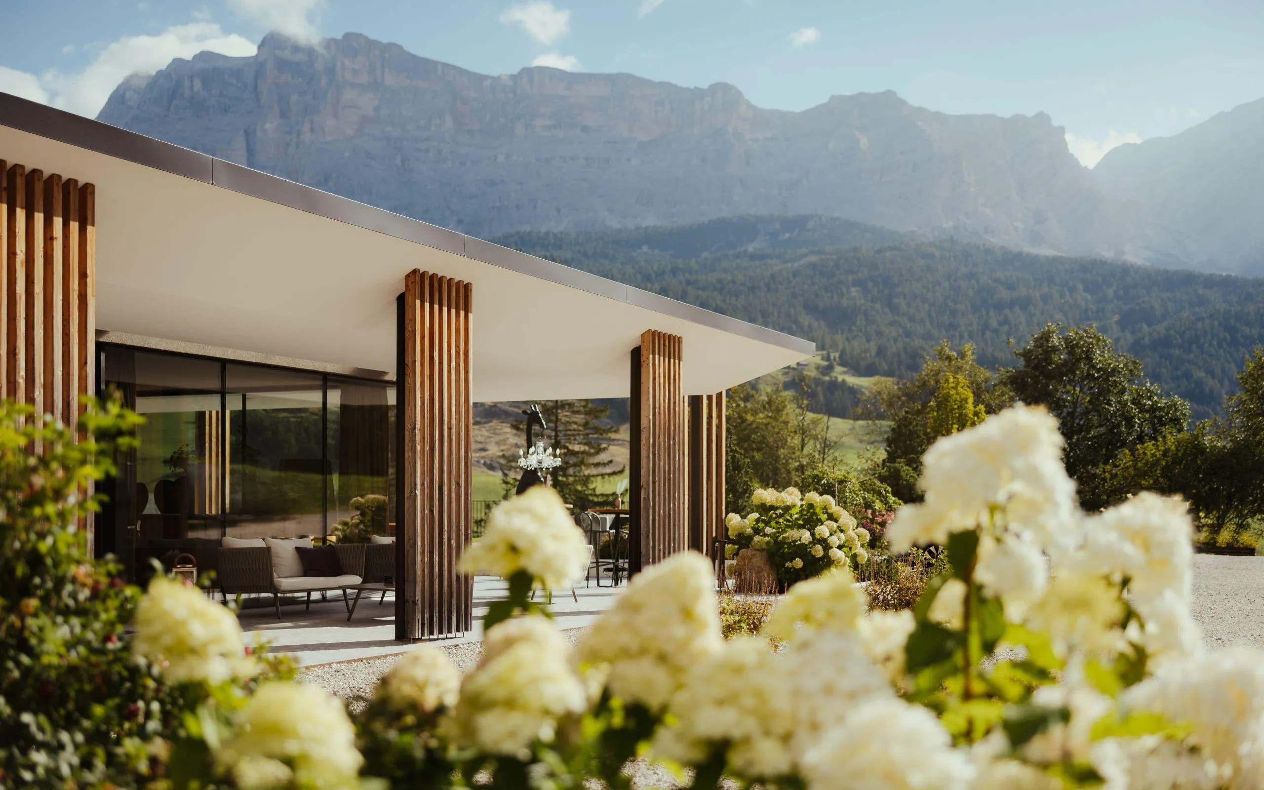 A modern wooden facade of Hotel Lech da Sompunt in La Villa, Alta Badia, is framed by lush white flowers and stunning Dolomites mountains.