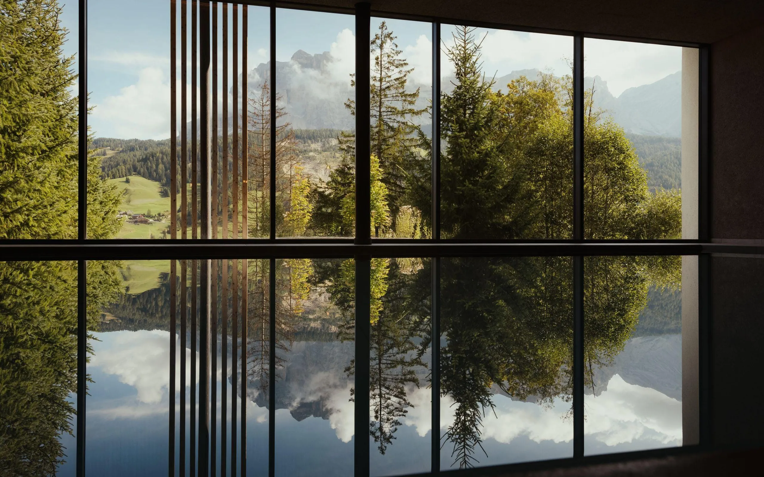 La vista serena dall'Hotel Lech da Sompunt mette in mostra alberi rigogliosi e maestose vette dolomitiche riflesse nell'acqua calma, incorniciate da moderne vetrate.