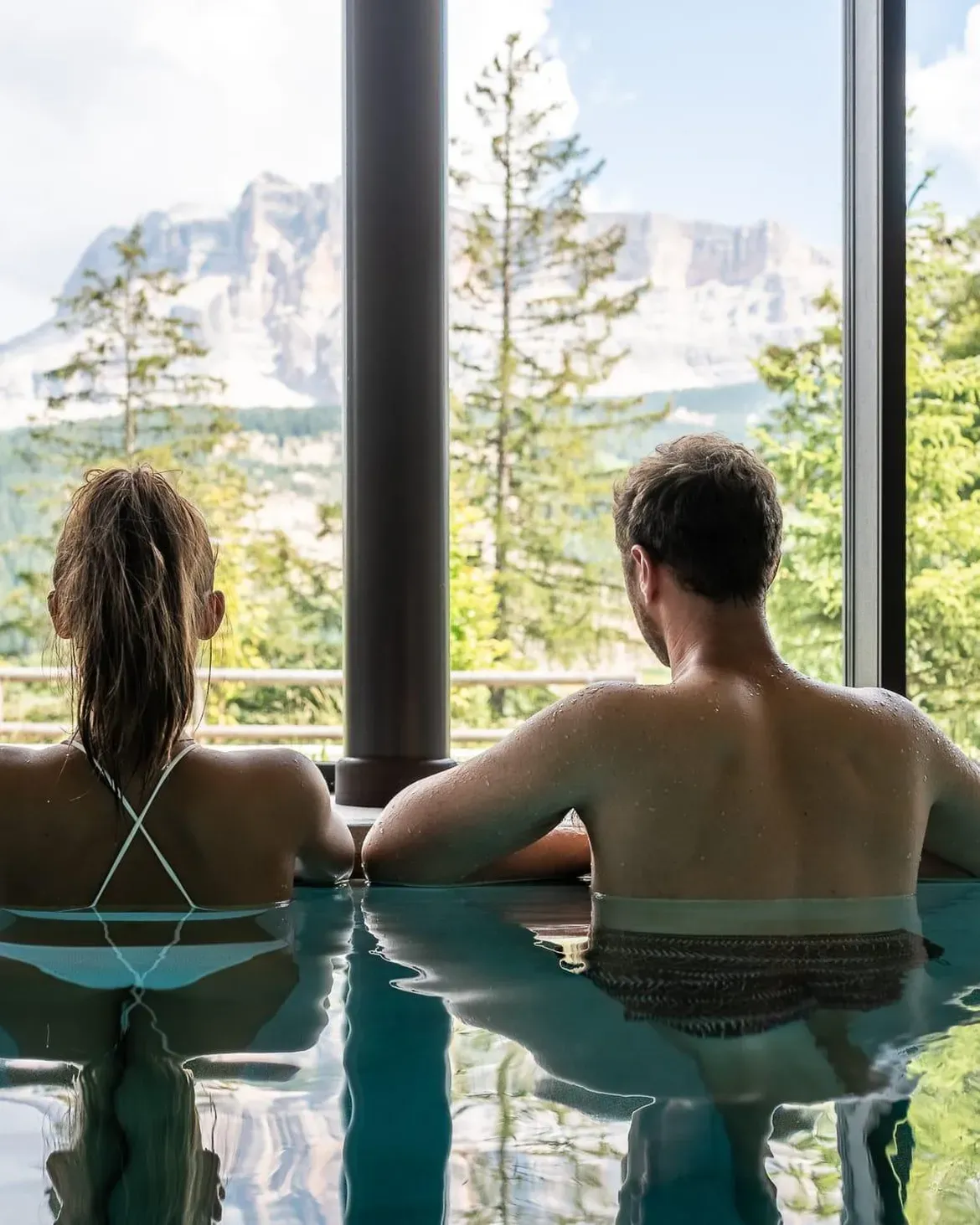 Una coppia si rilassa nella tranquilla piscina coperta dell'Hotel Lech da Sompunt, con vista sulle splendide Dolomiti e sulla vegetazione lussureggiante.