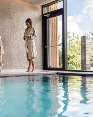 Una donna in accappatoio beige è in piedi accanto alla piscina coperta dell'Hotel Lech da Sompunt in Alta Badia.