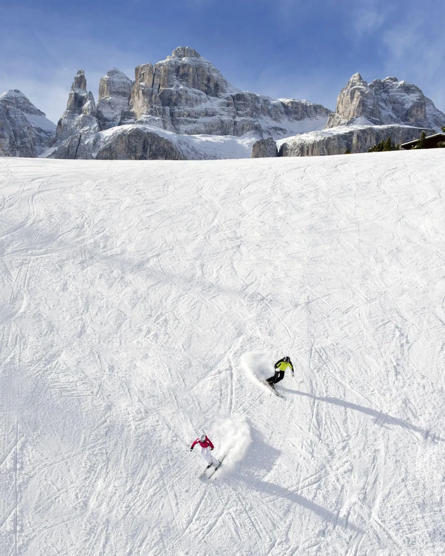 Two skiers carve through pristine snow in Alta Badia, casting long shadows on the slope.