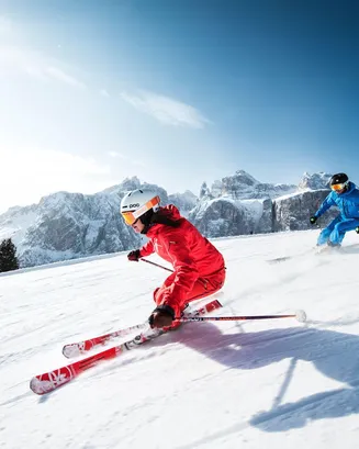 Due sciatori scendono a tutta velocità lungo un pendio innevato, sullo sfondo delle aspre montagne delle Dolomiti.