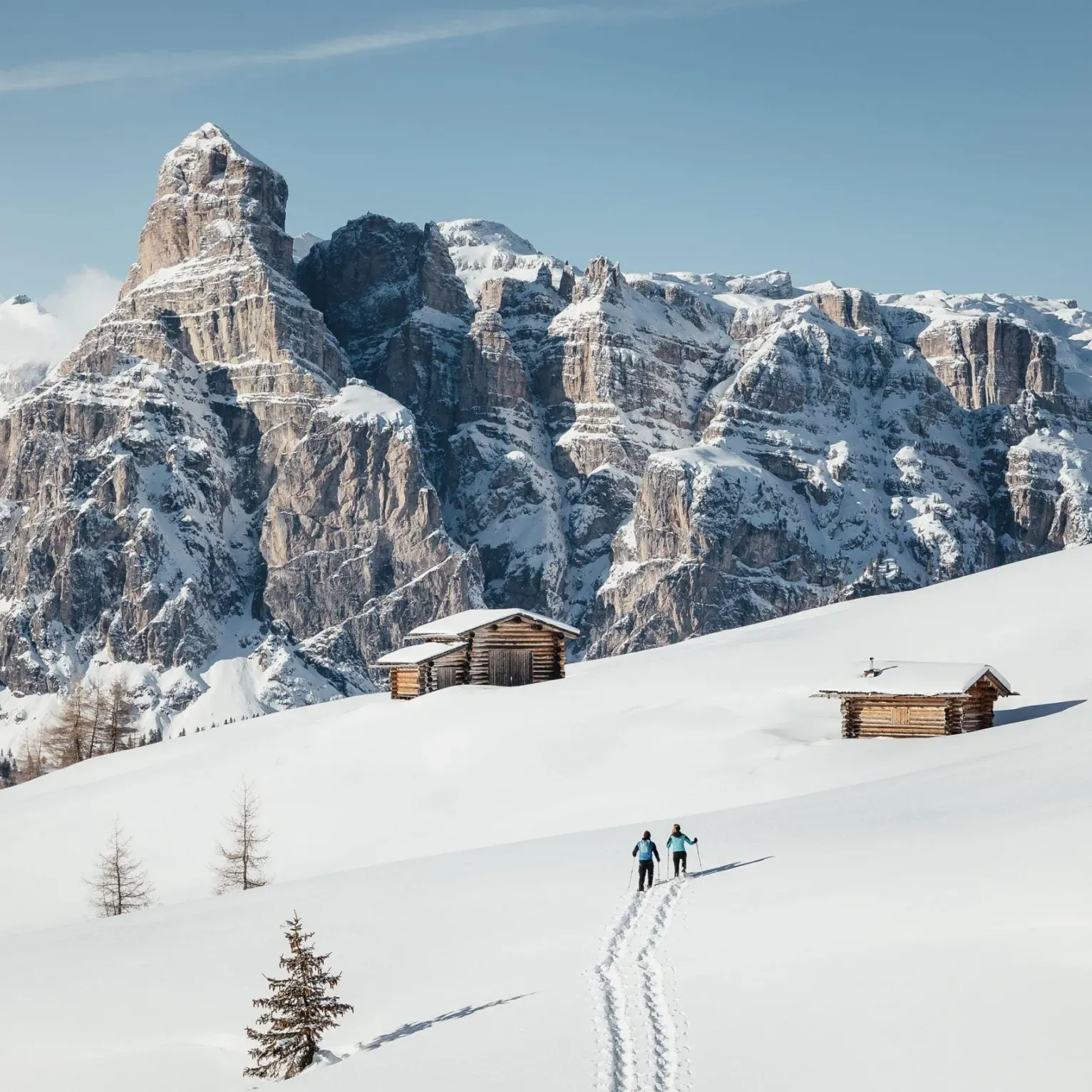 Two people snowshoeing through a snowy landscape towards rustic wooden huts, framed by the imposing snow-capped Sassolungo in the Dolomites, under a clear blue sky.