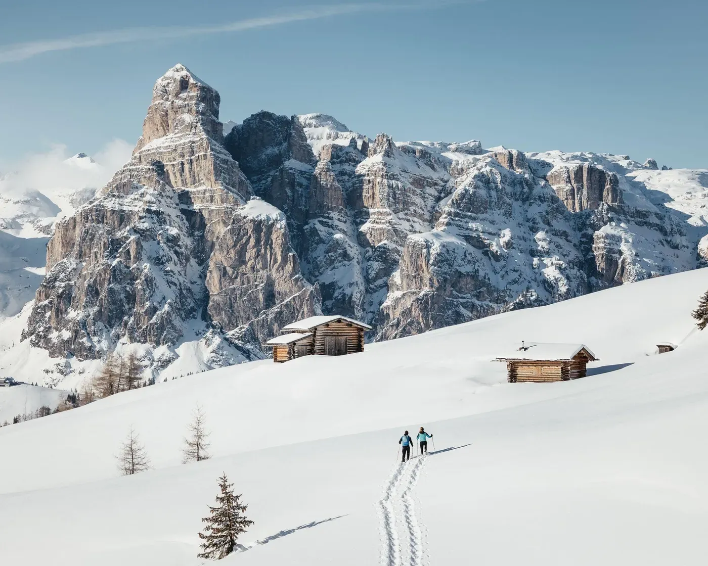 Due persone che ciaspolano attraverso un paesaggio innevato verso rustiche baite di legno, incorniciate dall'imponente Sassolungo innevato nelle Dolomiti, sotto un limpido cielo azzurro.