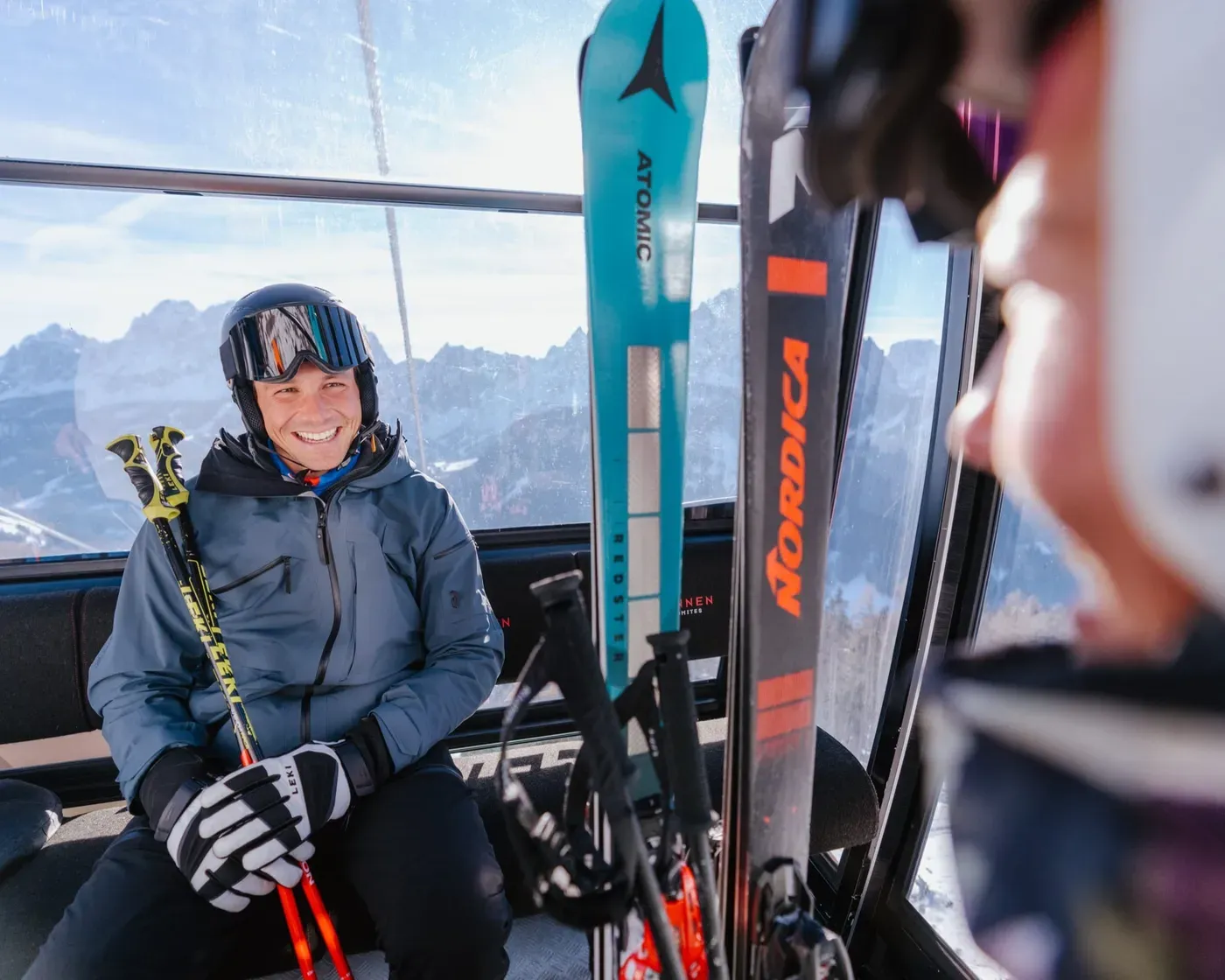 Uno sciatore sorridente seduto in una cabinovia, con in mano i bastoncini da sci.