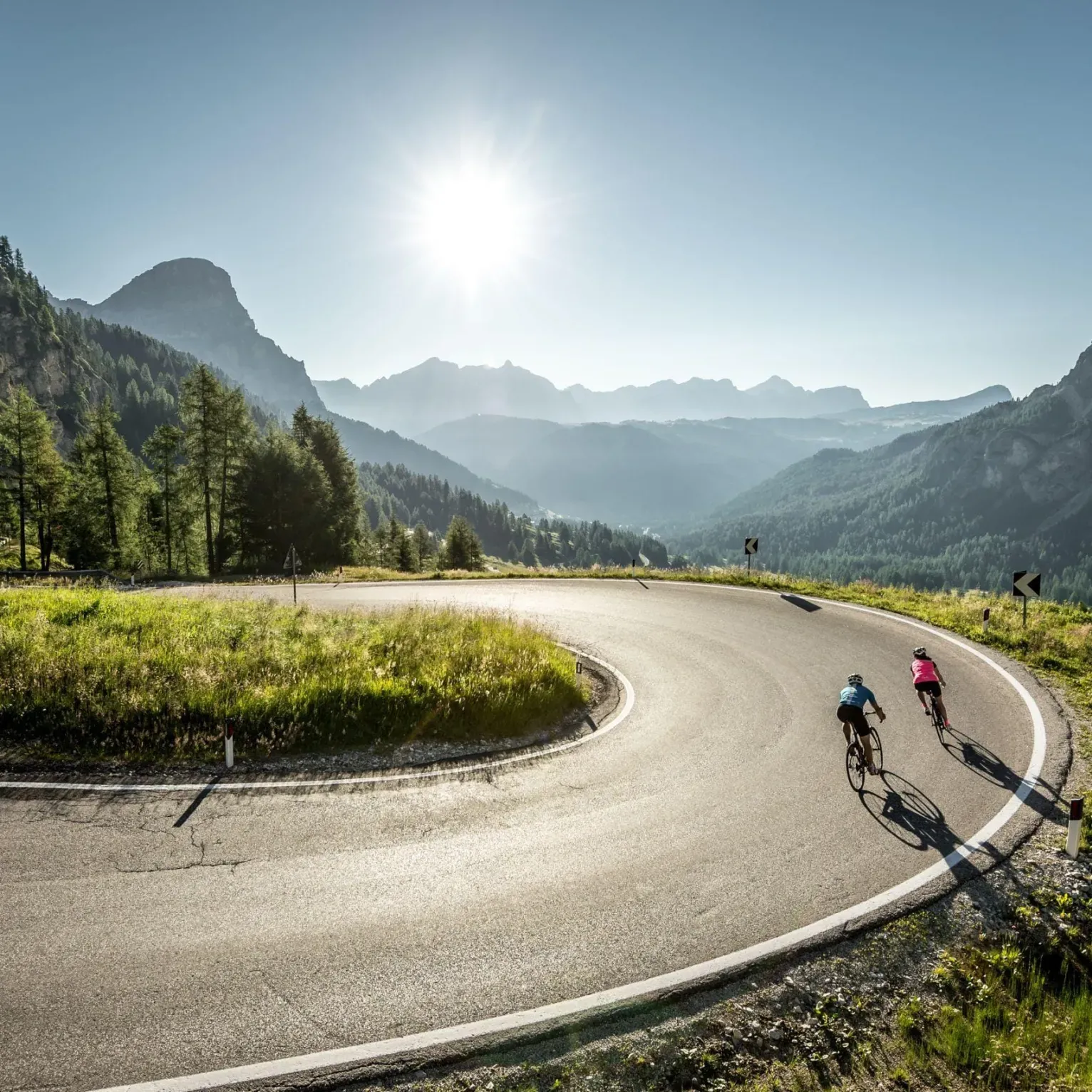 Due ciclisti percorrono la strada tortuosa del Passo Gardena in Alta Badia, attraversando un paesaggio montuoso e illuminati dalla calda luce del sole.