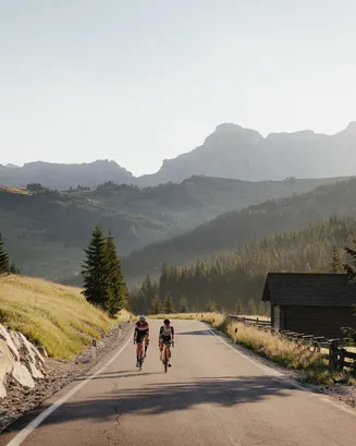 Due ciclisti percorrono la strada tranquilla del Passo Campolongo del Sellaronda, incorniciati da montagne e conifere nella morbida luce del mattino. Nelle vicinanze si può vedere una pittoresca baita.