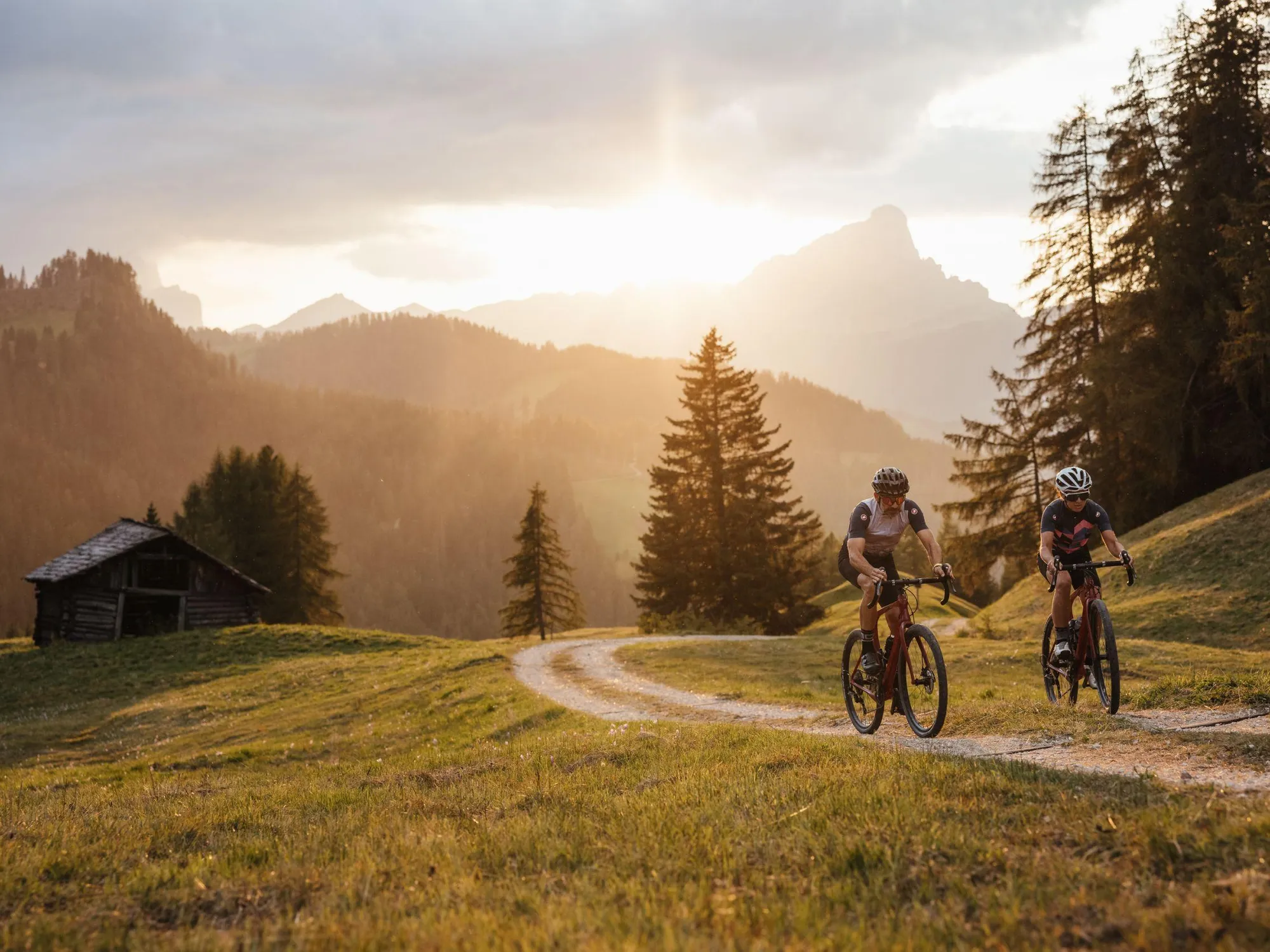 Due ciclisti percorrono un tortuoso sentiero sterrato a La Val, in Alta Badia, con una baita in legno e le Dolomiti sullo sfondo, sotto un cielo al tramonto.