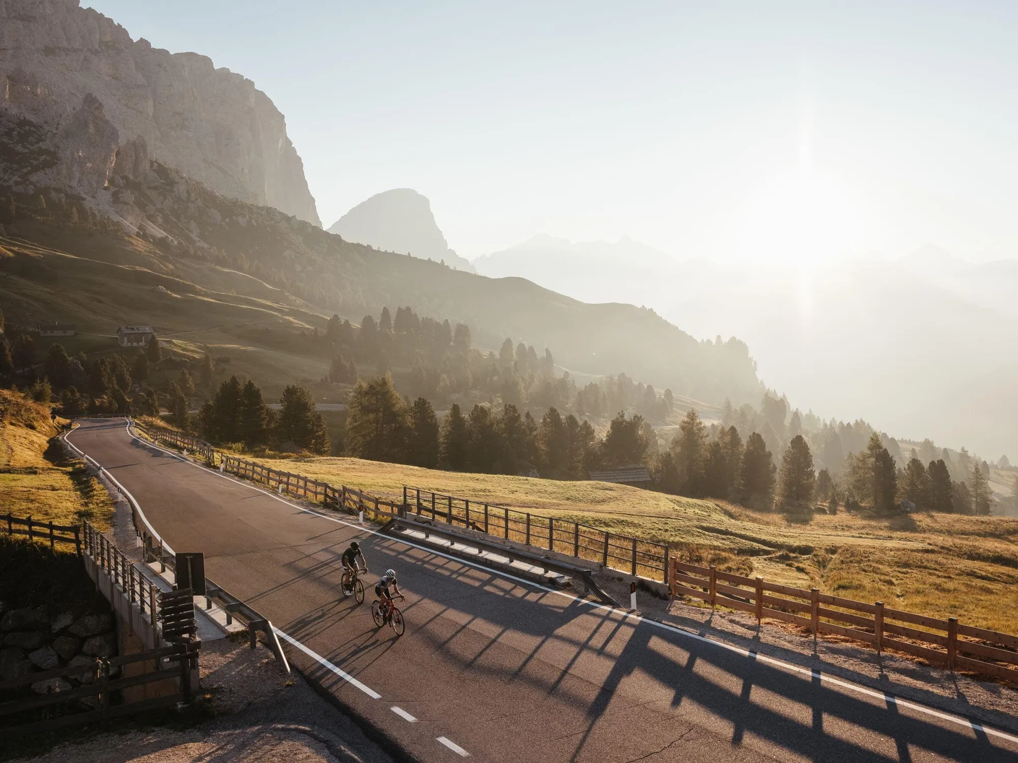 Due ciclisti percorrono la strada tortuosa del Passo Gardena sul Sellaronda, immersi in un sereno paesaggio dolomitico all'alba, con cime illuminate dal sole e alberi sullo sfondo.