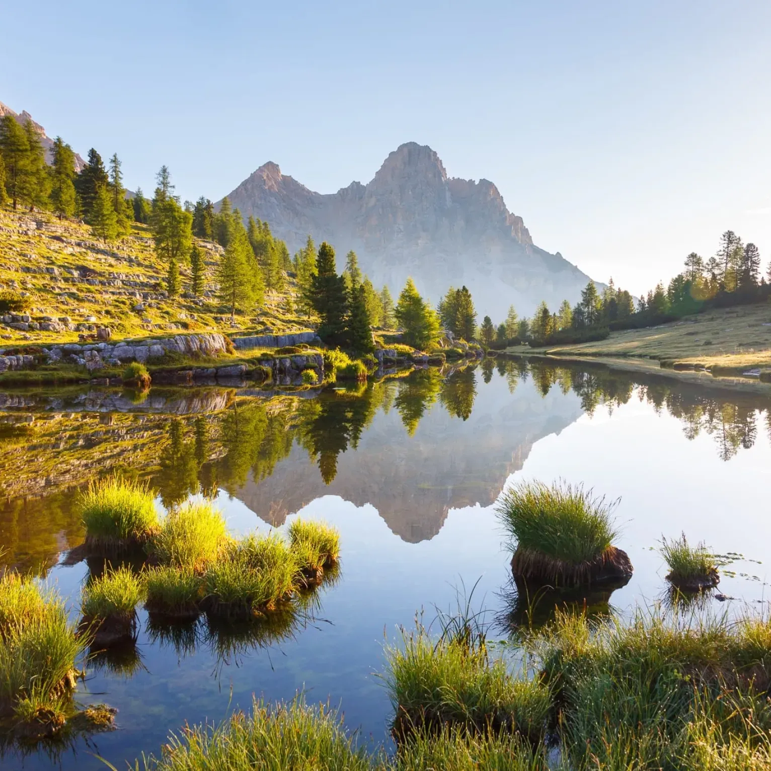 Il lago di Lé Vërt nel parco naturale Fanes-Senes-Braies riflette le imponenti montagne, circondato da vegetazione lussureggiante e rocce. La luce del sole mattutino crea una scena tranquilla e pittoresca.