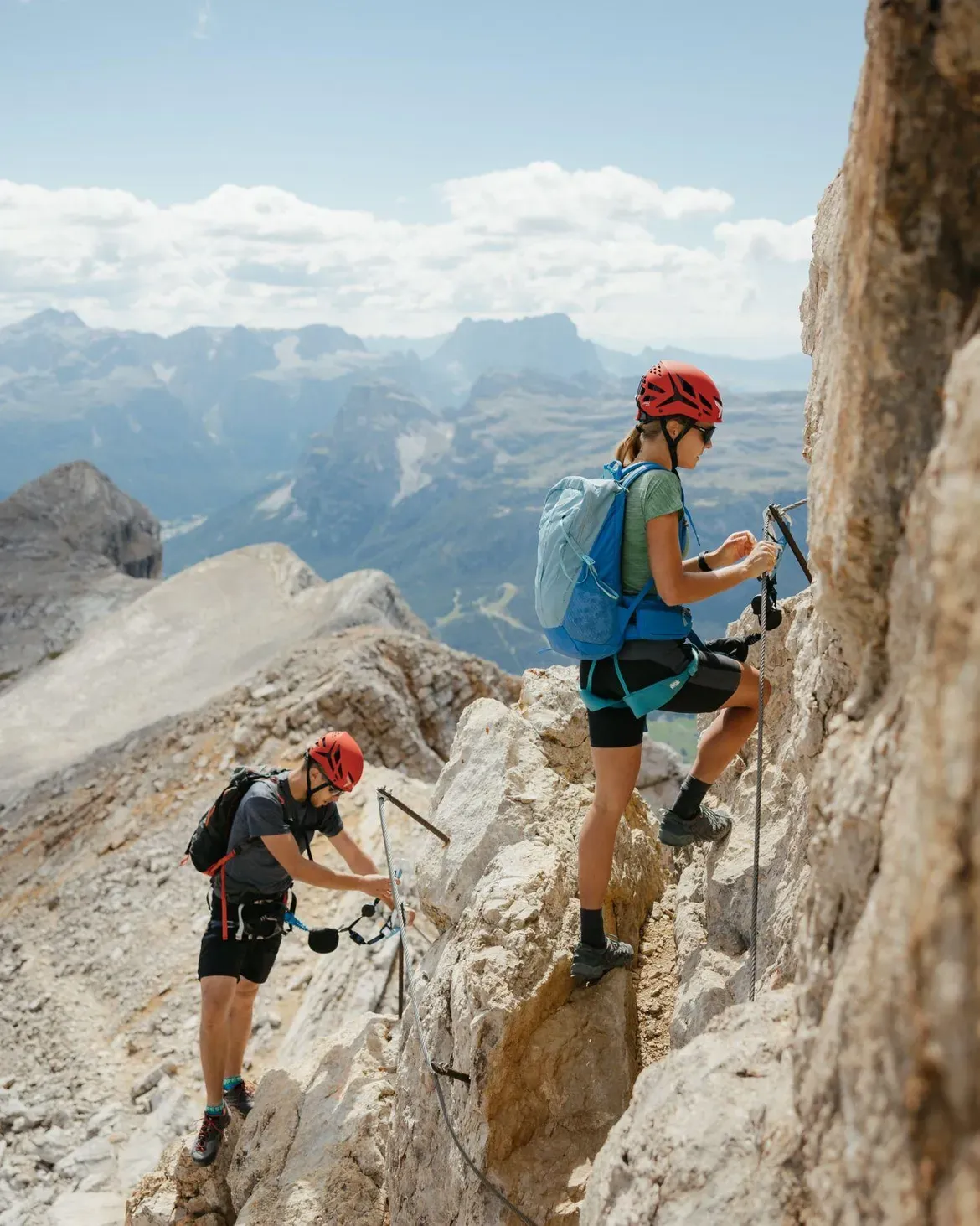 Due scalatori percorrono un sentiero roccioso sulla Cima Dieci in Alta Badia, dotati di casco e imbracatura, con l'ampio scenario delle Dolomiti sullo sfondo.