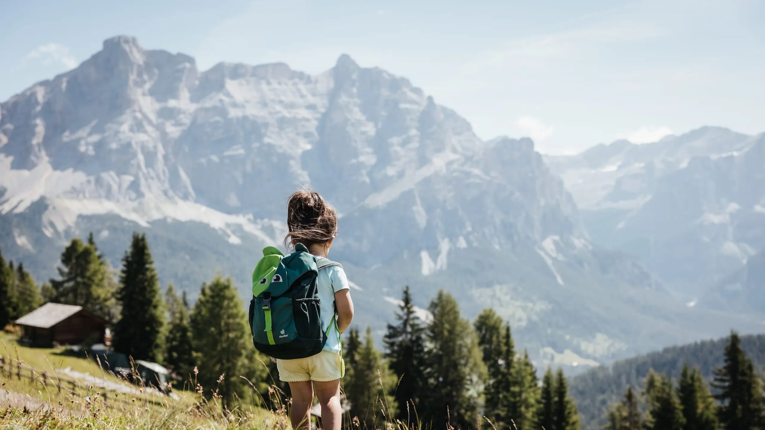 Un bambino con uno zaino è in piedi su un prato e ammira la catena del Sas dla Crusc nelle Dolomiti.