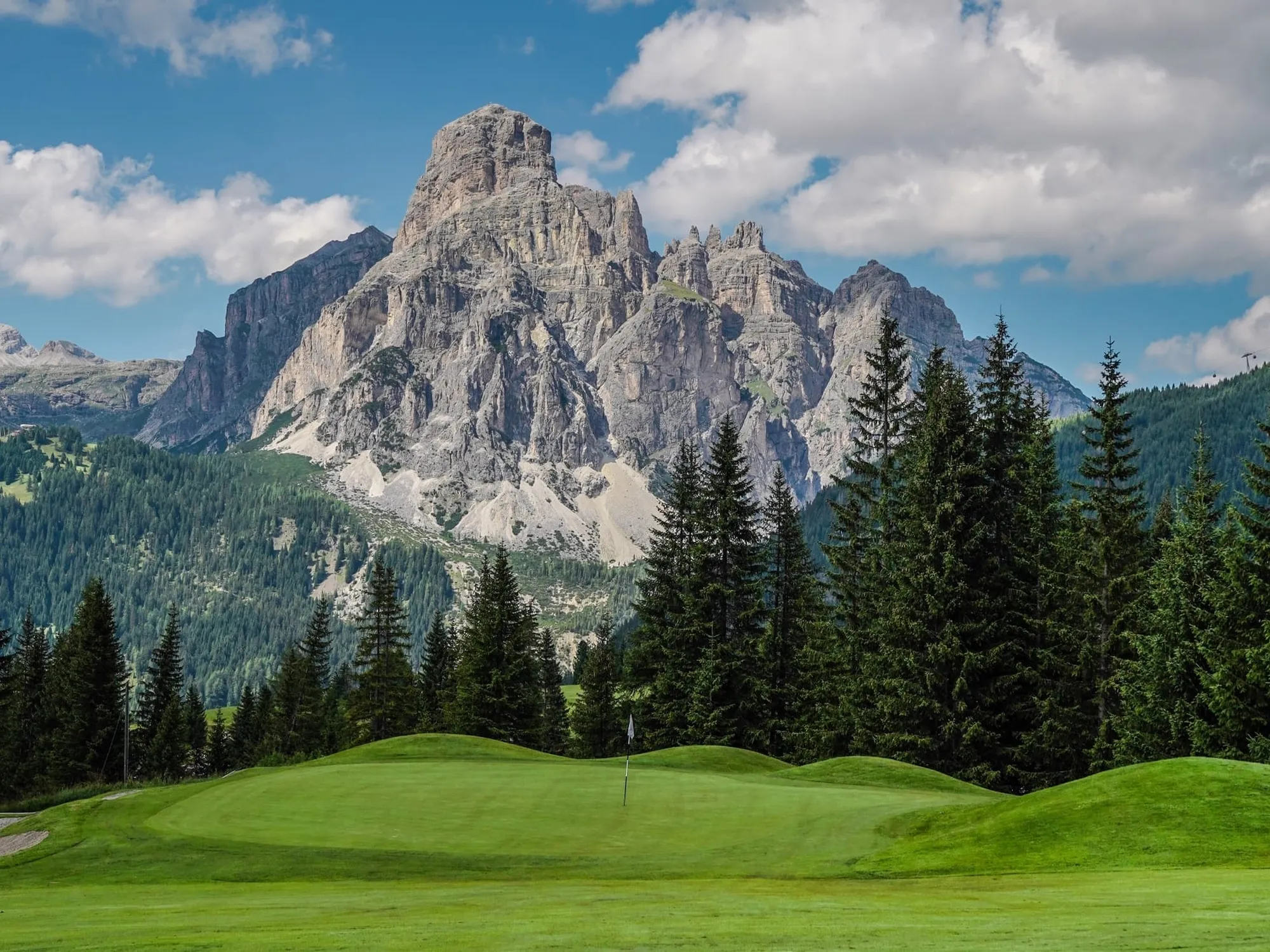 Centro golfistico di Corvara. Colline verdeggianti e un campo da golf in primo piano, incorniciati da alti alberi sempreverdi, il massiccio del Sassongher sullo sfondo.