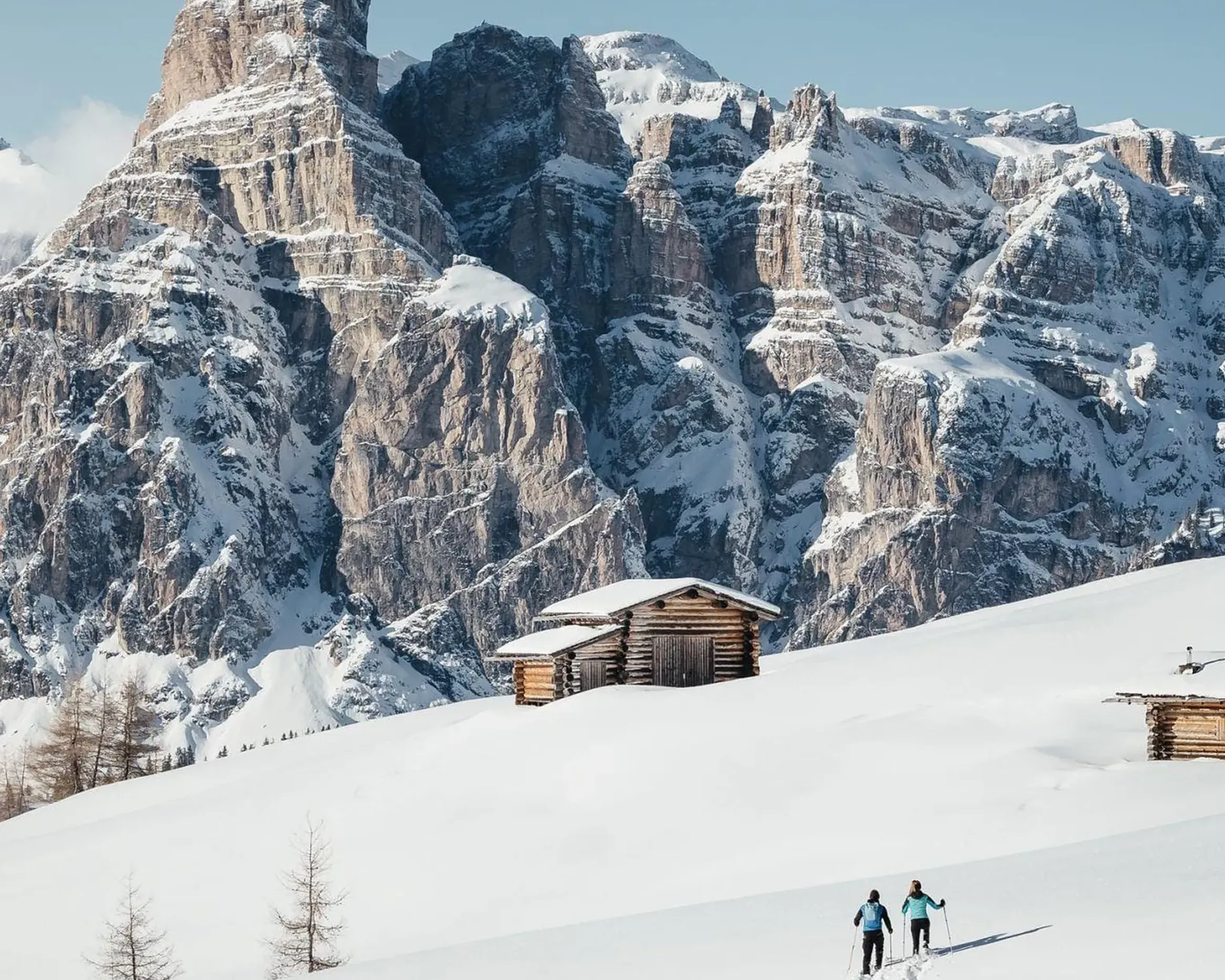 Due persone che ciaspolano attraverso un paesaggio innevato verso rustiche baite di legno, incorniciate dall'imponente Sassolungo innevato nelle Dolomiti, sotto un limpido cielo azzurro.