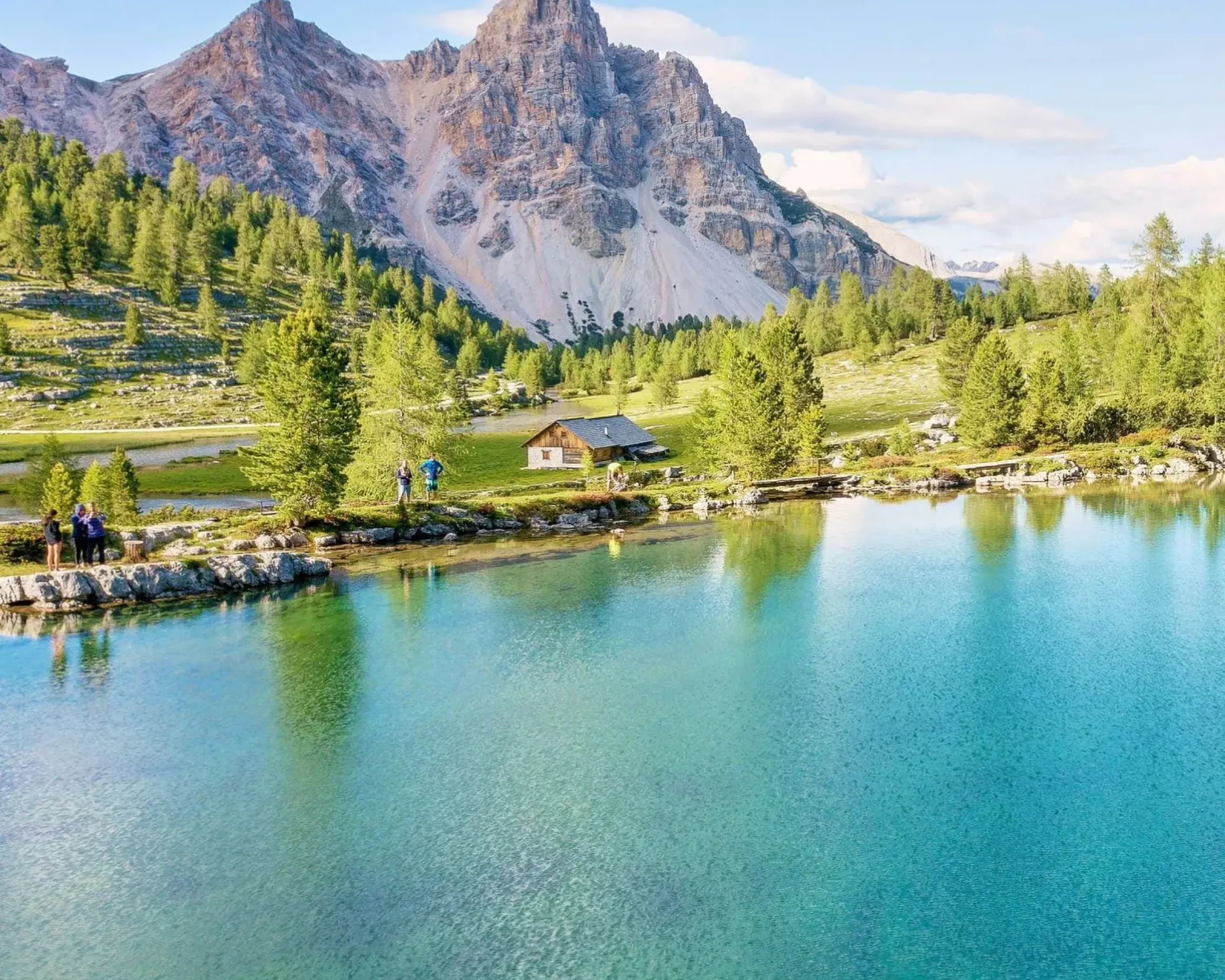 Il tranquillo lago alpino di Lé Vert, nel parco naturale Fanes-Senes-Braies, riflette le imponenti montagne e una vegetazione lussureggiante, con una piccola baita e persone che si godono il paesaggio lungo la riva.