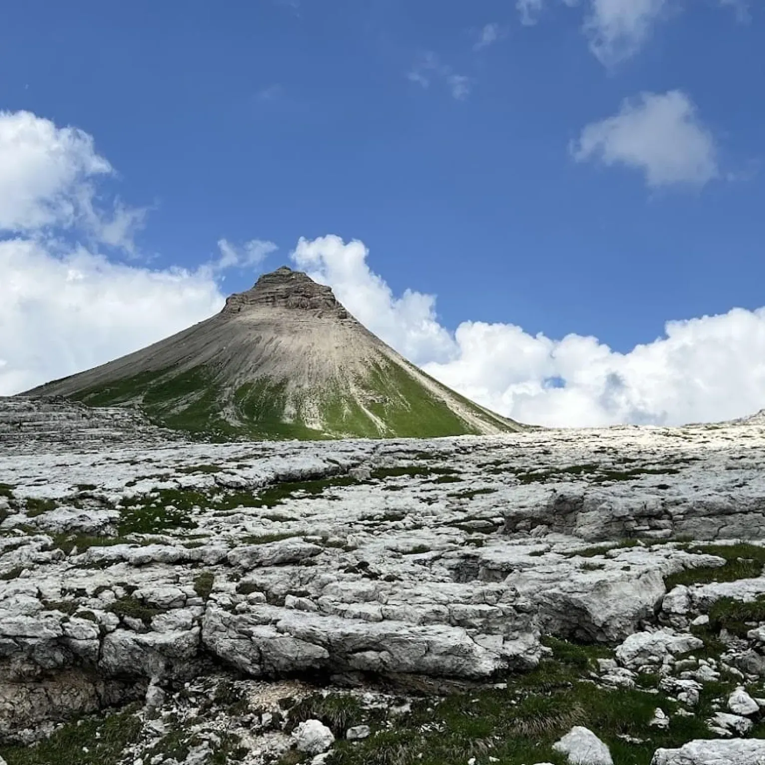 La cima frastagliata del Col dala Sune, nel parco naturale Puez-Odle, si staglia contro un cielo azzurro brillante ornato da soffici nuvole, circondata da un terreno roccioso e da distese di erba verde.