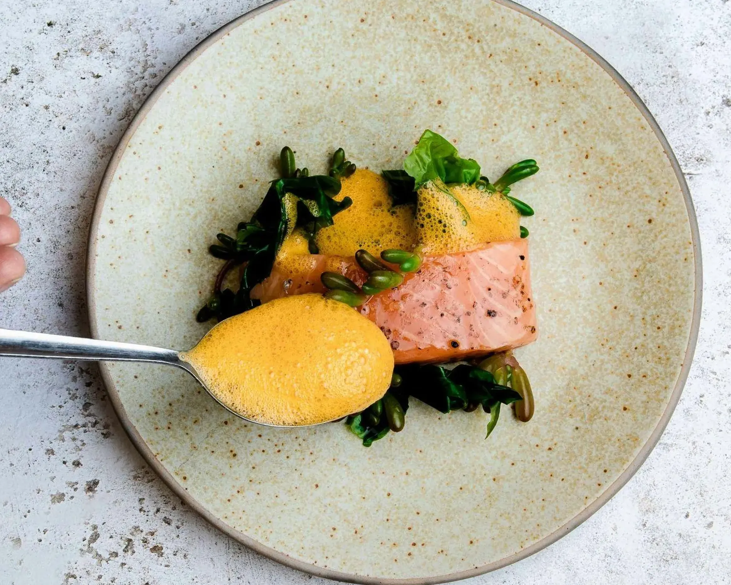 A hand holding a spoon with a creamy sauce over a plate of salmon and greens, set against a textured surface in Hotel Lech da Sompunt.