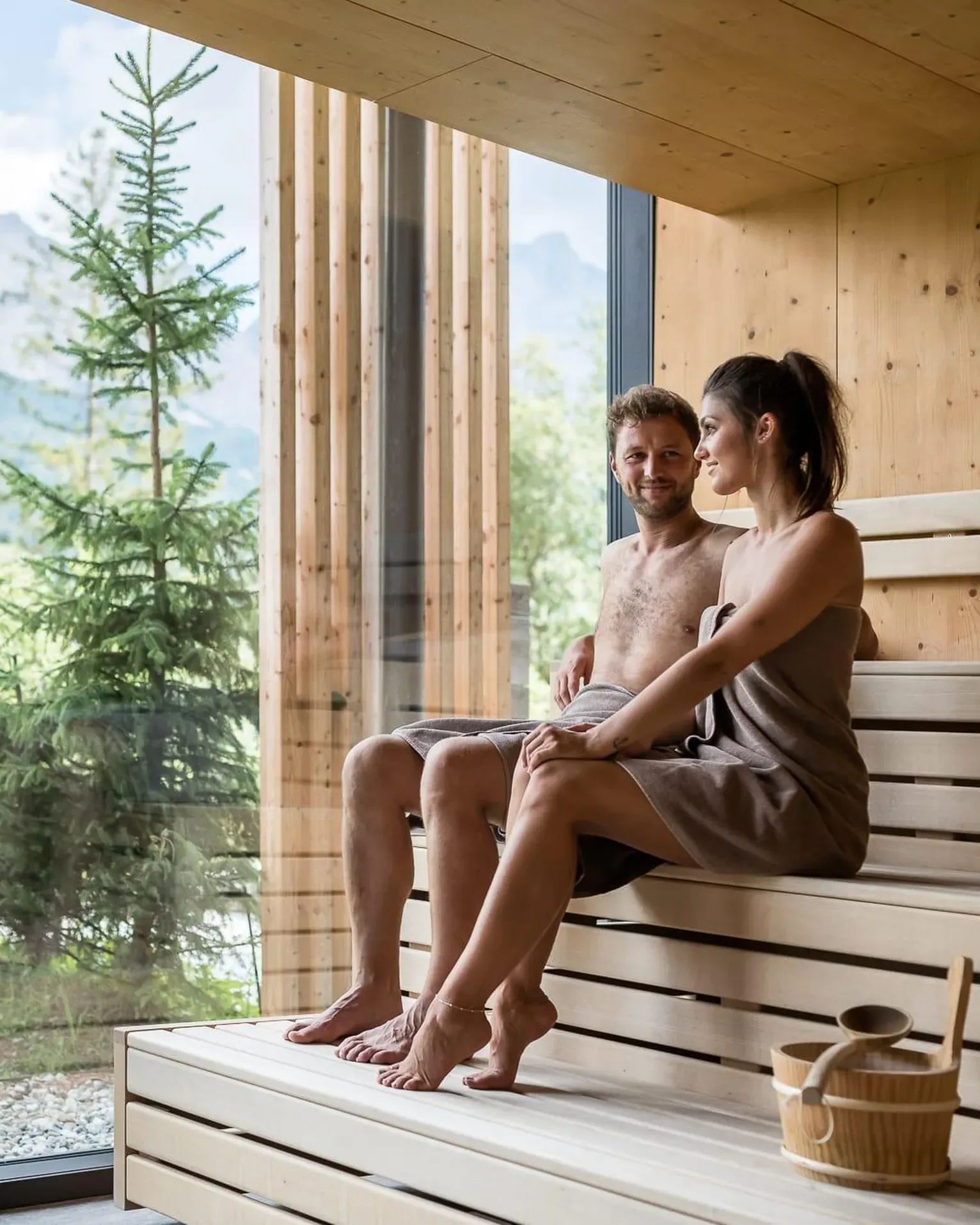 A man and woman sit on wooden benches in a sauna, wrapped in towels, enjoying a conversation with a view of trees and mountains outside.