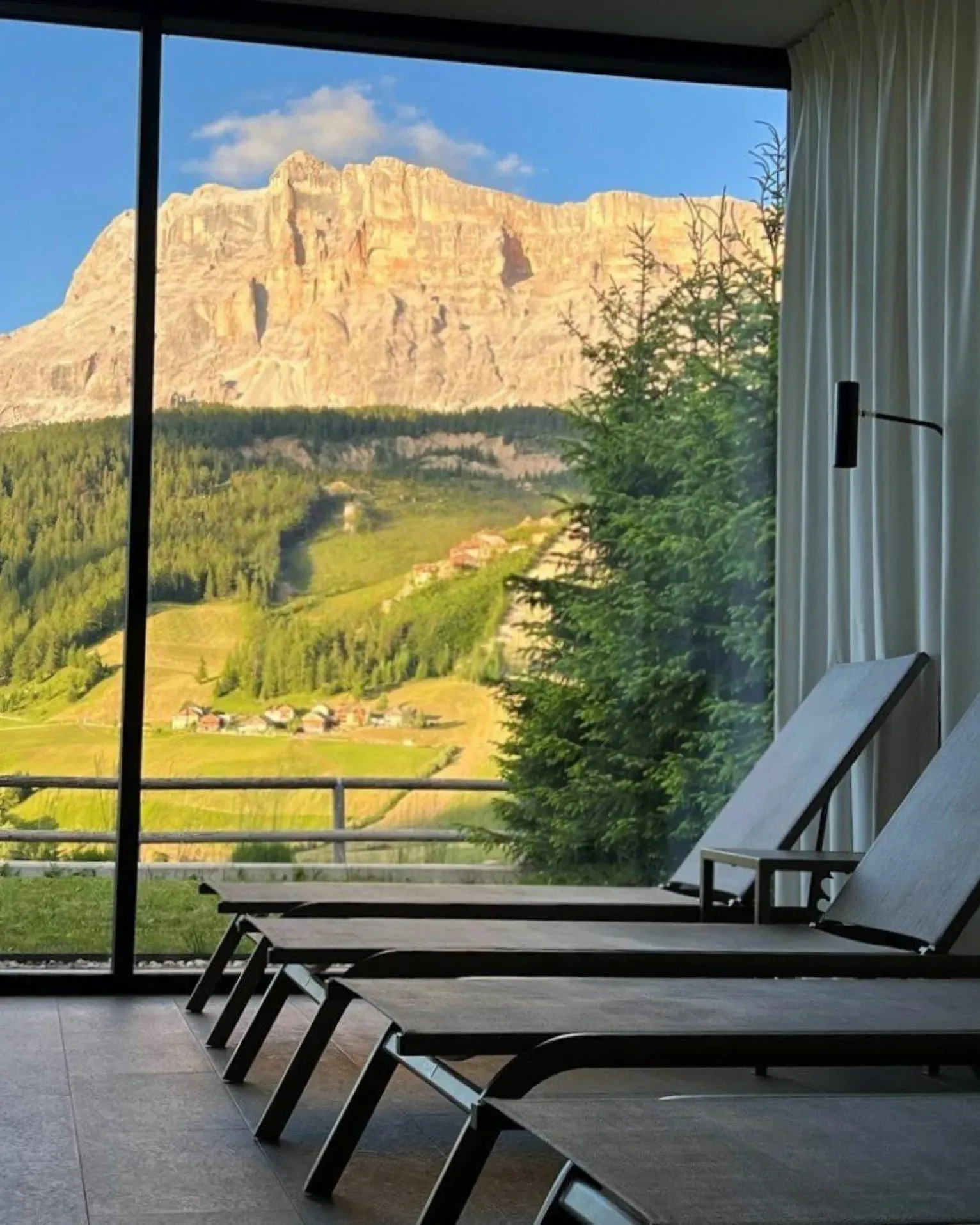 A serene relaxation room with three lounge chairs facing a large window, showcasing the Sas dla Crusc in Alta Badia view framed by lush greenery and clear skies.